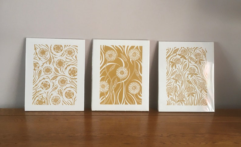 May include: Three framed prints with a white background and gold floral designs. The prints feature different floral patterns, including a repeating pattern of flowers and leaves, a pattern of stylized flowers with long stems, and a pattern of clover-like flowers.