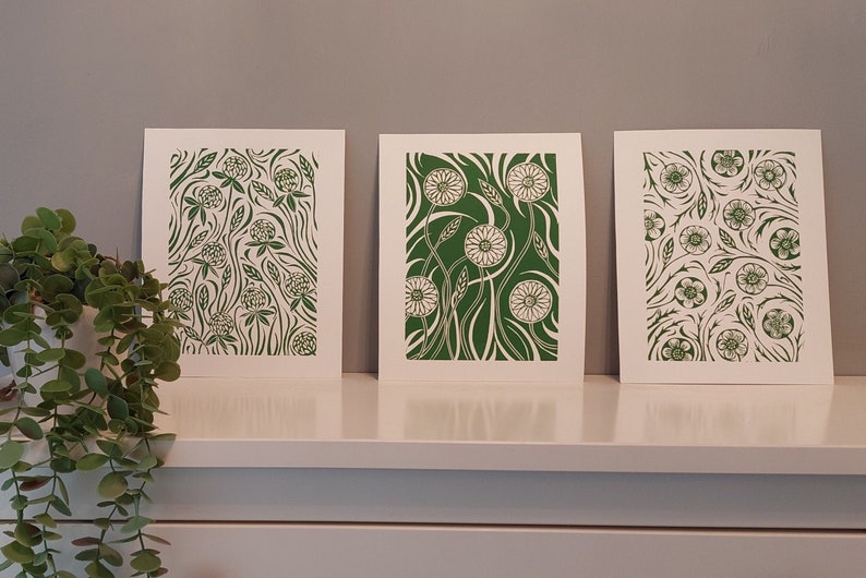 May include: Three framed prints with green floral designs. The prints feature different floral patterns, including clover, dandelions, and other flowers. The prints are all on white backgrounds.