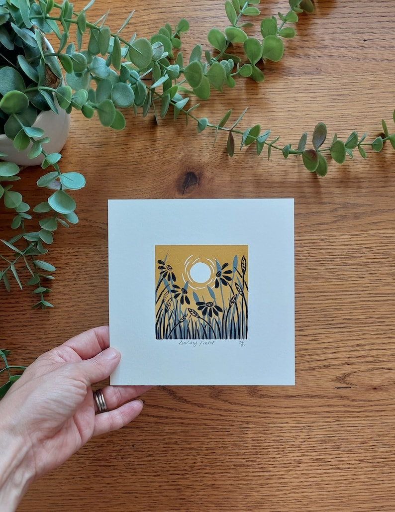 May include: A print of a yellow field with blue flowers and a white sun. The print is titled "Daisy Field" and signed by the artist.