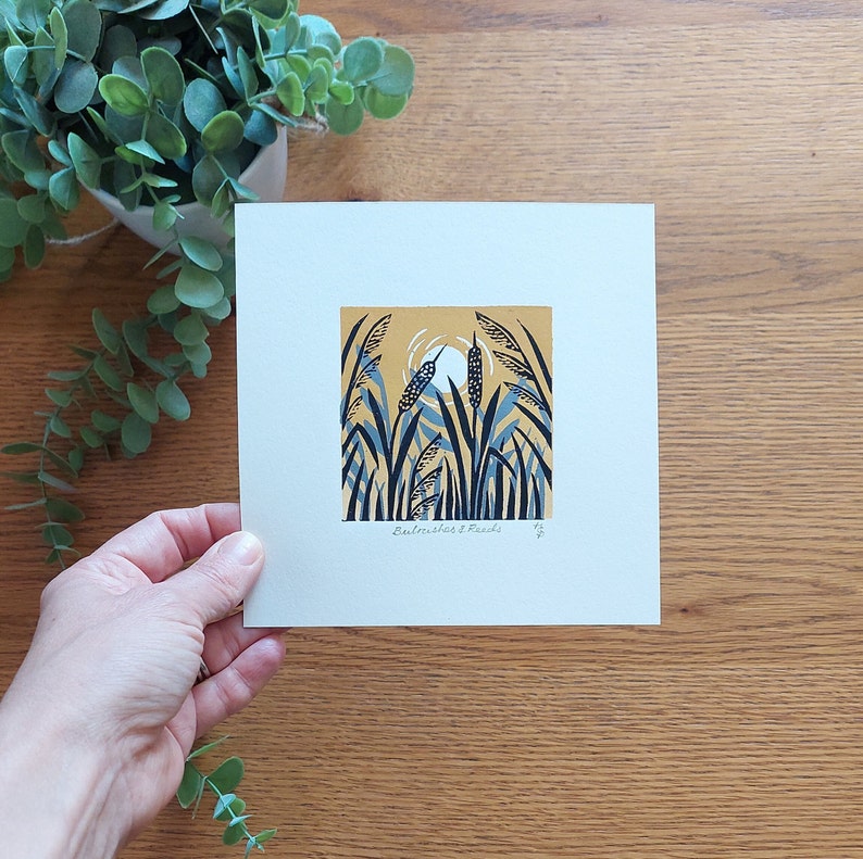 May include: A blue and yellow print of a stylised landscape with tall reeds and a bird in the centre. The print is on a white background and has the text "Buttercup & Finch" in the bottom centre.