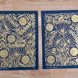 May include: Two floral art prints with gold foil designs on a navy blue background. The prints feature stylized flowers and foliage. The text "Thoughts of flowers" is visible on the bottom of one print. The prints are displayed on a wooden surface.