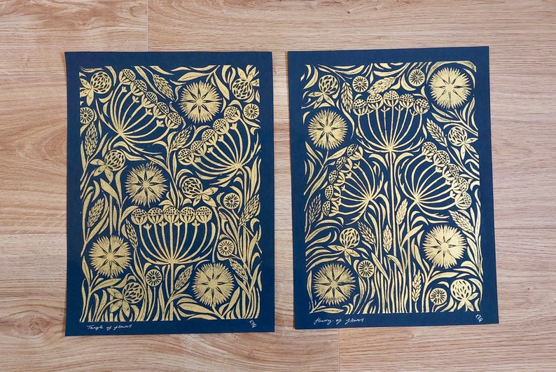 May include: Two floral art prints with gold foil designs on a navy blue background. The prints feature stylized flowers and foliage. The text "Thoughts of flowers" is visible on the bottom of one print. The prints are displayed on a wooden surface.