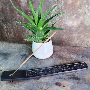 Puede incluir: Un porta incienso negro con un diseño intrincado y un palo de incienso de madera descansando sobre él. Una planta suculenta verde en una maceta blanca está en el fondo.