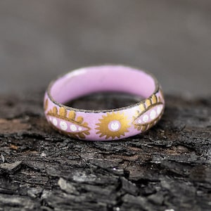 May include: A pink ring with a gold and white floral design. The ring is on a dark brown wooden surface.