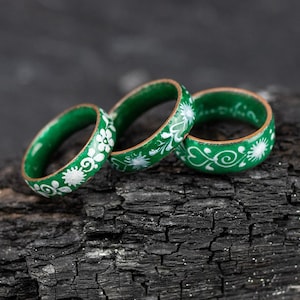 May include: Three green wooden rings with white floral designs. The rings are stacked on top of each other on a black, charred surface.