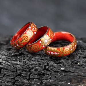 May include: Three red wooden rings with gold and white painted floral designs. The rings are stacked on top of each other on a black charred wooden surface.