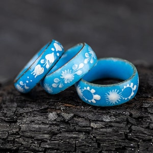 May include: Three blue rings with white floral designs stacked on top of each other. The rings are on a black, charred wooden surface.