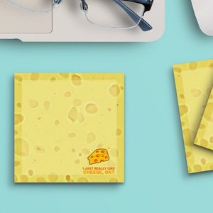 May include: Two yellow square notepads with a cheese pattern and the text "I just really like cheese, ok?" printed on them. The notepads are on a light blue surface with a laptop, glasses, a computer mouse, a cup of coffee, and paper clips.