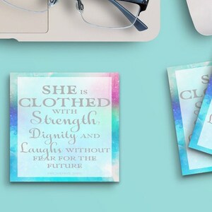 Puede incluir: Vista superior de un espacio de trabajo con un portátil, gafas, un ratón blanco y una taza de café. Dos posavasos cuadrados con el texto "SHE IS CLOTHED WITH Strength, Dignity AND Laughs WITHOUT FEAR FOR THE FUTURE."