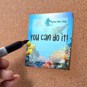 May include: A blue and white motivational sticky note with the text "Seas the day" and "You can do it!"  The note has a picture of a shark swimming in the ocean with coral reefs and fish.