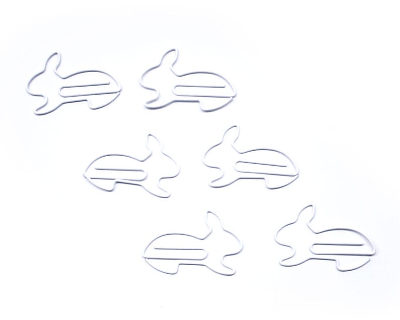May include: Six white metal bunny-shaped paper clips. The clips are arranged in a staggered pattern on a white background.