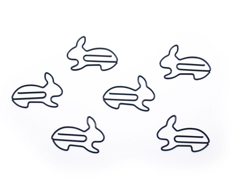 May include: Six black metal paper clips shaped like rabbits. The clips are arranged in a scattered pattern on a white background.