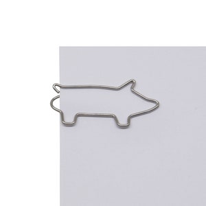 May include: Silver metal pig-shaped paperclip. The paperclip is holding a sheet of white paper.
