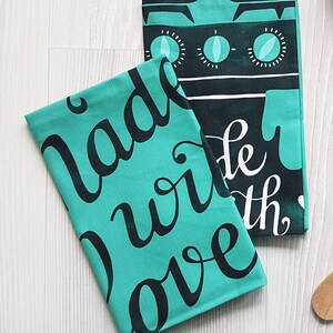 May include: Two teal kitchen towels with black text. One towel reads "Made with Love" in cursive. The other towel has a graphic of a stove top with the words "Made with" in white cursive. The towels are on a white wooden surface.