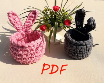 May include: Two crocheted bunny baskets, one pink and one gray, with a small plant in a white pot in the background. The text "PDF" is written in red on the white surface.