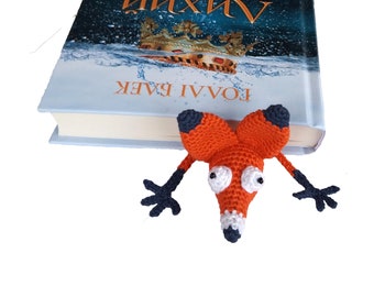 May include: A crocheted orange fox bookmark with black arms and white eyes. The bookmark is hanging off the edge of a book titled "Лихий Король" by Голлі Блек.