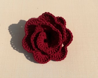 May include: A deep red crocheted flower with multiple layers. The flower is made with a single strand of yarn and has a textured, slightly bumpy surface.