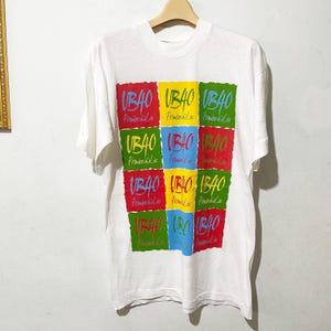 Puede incluir: Camiseta blanca con un diseño gráfico colorido. El diseño presenta el texto "UB40" en una cuadrícula de cuadrados coloridos. La camiseta está colgada en una percha de madera.