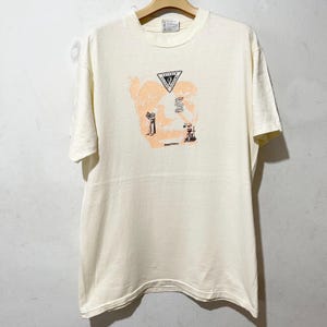 May include: Cream-colored t-shirt with a graphic design. The design features a triangle with text, a stylized figure, and the word "Raychem" in a sans-serif font. The shirt is hanging on a wooden hanger.