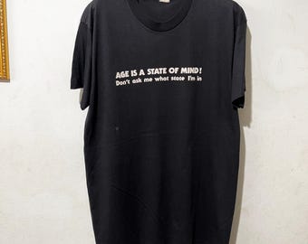 May include: Black t-shirt with white text that reads "AGE IS A STATE OF MIND! Don't ask me what state I'm in." The shirt has short sleeves and a crew neck. The shirt is hanging on a wooden hanger.