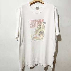 Könnte beinhalten: Weißes T-Shirt mit Rundhalsausschnitt und kurzen Ärmeln. Auf der Vorderseite befindet sich eine verblasste Grafik mit dem Text "LA BALLADE DE LA MER SALEE" und Illustrationen von Männern mit Militärmützen. Das Shirt hängt an einem Holzkleiderbügel.