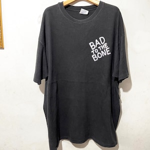 May include: Black t-shirt with the phrase "BAD TO THE BONE" printed in white on the front. The shirt is hanging on a wooden hanger. The t-shirt has short sleeves and a crew neck.