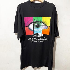 May include: Black t-shirt with a graphic eye design. The eye is split into four coloured sections: pink, green, yellow, and blue. The text "Serif SPC" and the slogan "Keeping an Eye on the Best Designers in the World" are below the eye.