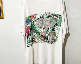 May include: White t-shirt featuring a colorful jungle scene with an elephant, giraffe, zebra, leopard, parrot, and deer. The graphic is set against a backdrop of lush green foliage. The shirt is hanging on a wooden hanger.