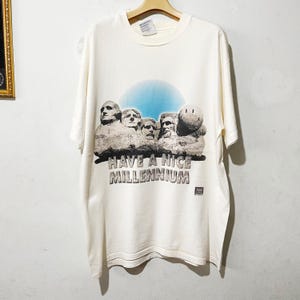 Puede incluir: Camiseta de color crema con un gráfico del Monte Rushmore, una carita sonriente y el texto "HAVE A NICE MILLENNIUM" en gris. El diseño incluye un fondo de cielo azul. La camiseta está colgada en una percha de madera.