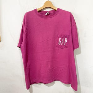 May include: A short-sleeved magenta t-shirt with a crew neckline and a single chest pocket on the left side. The word GAP is printed in white capital letters across the front of the pocket. The shirt is displayed on a wooden hanger.