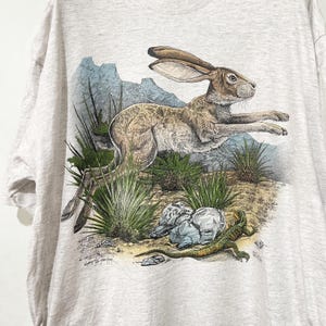 May include: A light gray t-shirt featuring a detailed illustration of a running jackrabbit. The design includes desert plants, rocks, and a lizard, with a backdrop of blue mountains. The artwork is in a realistic style.