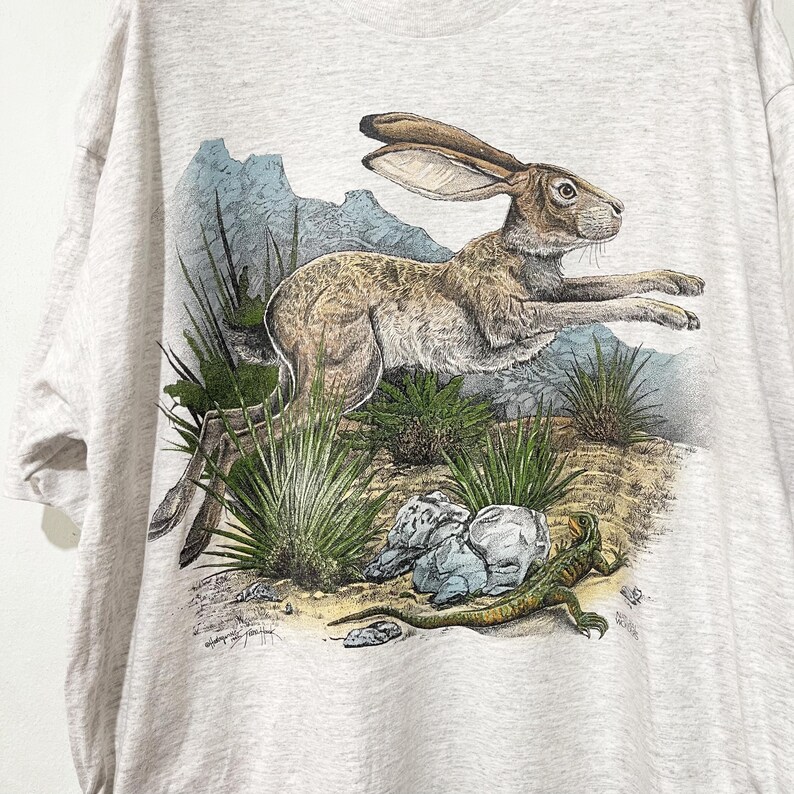 May include: A light gray t-shirt featuring a detailed illustration of a running jackrabbit. The design includes desert plants, rocks, and a lizard, with a backdrop of blue mountains. The artwork is in a realistic style.
