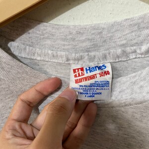 Puede incluir: Primer plano de una etiqueta de camiseta gris Hanes Heavyweight 50/50. La etiqueta es roja y blanca, con texto que indica que la camiseta est&aacute; hecha de 50% algod&oacute;n y 50% poli&eacute;ster. Talla X-Large.