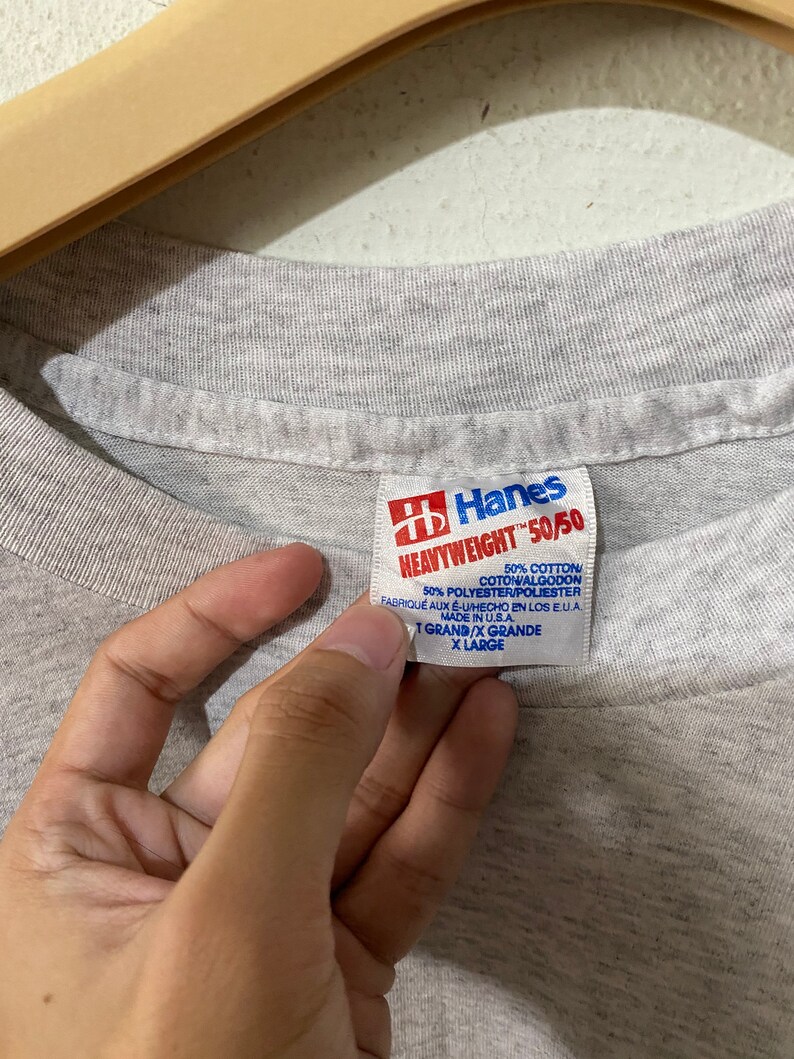 Puede incluir: Primer plano de una etiqueta de camiseta gris Hanes Heavyweight 50/50. La etiqueta es roja y blanca, con texto que indica que la camiseta est&aacute; hecha de 50% algod&oacute;n y 50% poli&eacute;ster. Talla X-Large.