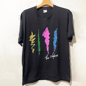May include: Black t-shirt with a colorful abstract design featuring yellow, green, pink, and blue vertical shapes. The text "The Nylons" is written in white script below the design. The shirt is hanging on a wooden hanger.