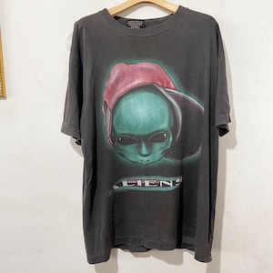 May include: Dark grey t-shirt with a green alien wearing a red hat. The word "ALIENS" is printed in white on a green banner below the alien's face. The shirt is displayed on a wooden hanger.