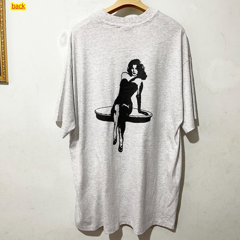 Puede incluir: Camiseta gris jaspeado con un gr&aacute;fico negro de una mujer sentada en una plataforma redonda. La mujer lleva un vestido sin tirantes y tacones. La camiseta tiene mangas cortas y cuello redondo.