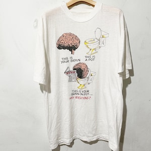 May include: White t-shirt with a humorous graphic. The design includes a detailed illustration of a brain, a toilet, and a brain on a toilet, with the text "THIS IS YOUR BRAIN", "THIS IS A POT", and "THIS IS YOUR BRAIN ON POT... ANY QUESTIONS?"