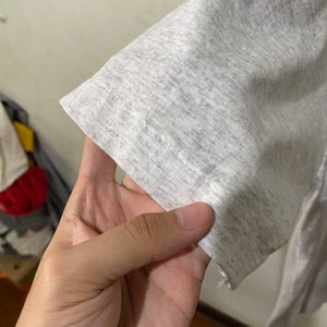 Puede incluir: Primer plano de una manga de camiseta gris claro. La tela tiene una textura jaspeada, con sutiles variaciones de color. El borde de la manga es visible, mostrando el dobladillo y los detalles de costura. El aspecto general es informal y c&oacute;modo.