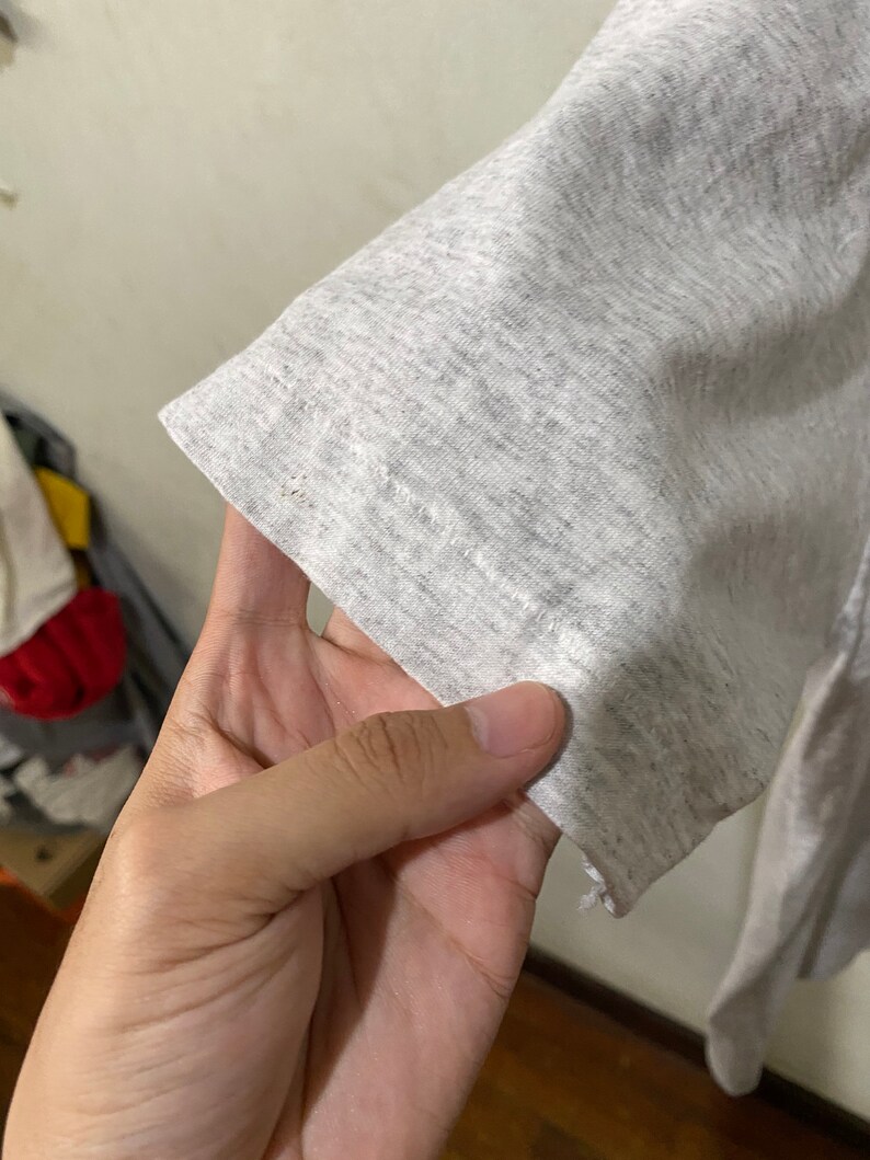 Puede incluir: Primer plano de una manga de camiseta gris claro. La tela tiene una textura jaspeada, con sutiles variaciones de color. El borde de la manga es visible, mostrando el dobladillo y los detalles de costura. El aspecto general es informal y c&oacute;modo.