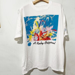 以下が含まれることがあります： 前面にカラフルな抽象的なデザインが施された白いTシャツ。デザインは、青い背景に赤、黄、青の飛沫が特徴です。「A Ruby Original」の文字が下に印刷されています。