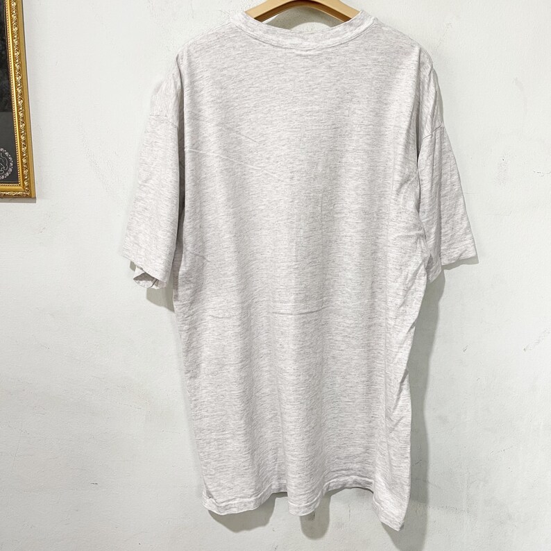May include: A heather gray t-shirt with short sleeves hangs on a wooden hanger. The shirt is made of a soft, textured fabric. The t-shirt is displayed against a white wall.