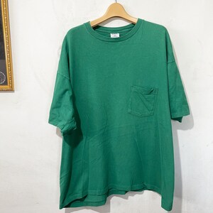 Könnte beinhalten: Ein einfarbiges grünes T-Shirt mit kurzen Ärmeln, Rundhalsausschnitt und einer aufgesetzten Tasche auf der linken Brustseite. Das Kleidungsstück hängt an einem Holzkleiderbügel und zeigt ein schlichtes Design mit geradem Saum.