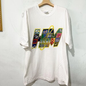 May include: White t-shirt with a colorful graphic design. The design features the letters "HM" with a treble clef, and several small images. The word "Hilo" is written in cursive. The shirt is hanging on a wooden hanger.