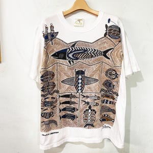 May include: White t-shirt with a brown and blue marine life design. The shirt features a large fish in the center, surrounded by other sea creatures. The words "HAWAIIAN DREAMING" are printed at the bottom. The shirt is hanging on a wooden hanger.