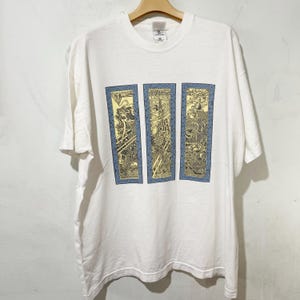 May include: White t-shirt with a crew neck and short sleeves. The shirt displays three vertical rectangular panels with detailed yellow and black designs on a blue backdrop. The t-shirt is hanging on a wooden hanger.