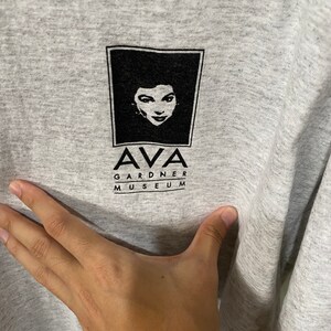 Puede incluir: Camiseta gris con un gr&aacute;fico en blanco y negro del rostro de una mujer dentro de un marco cuadrado. Debajo, el texto "AVA GARDNER MUSEUM" en negro, centrado en la camiseta.