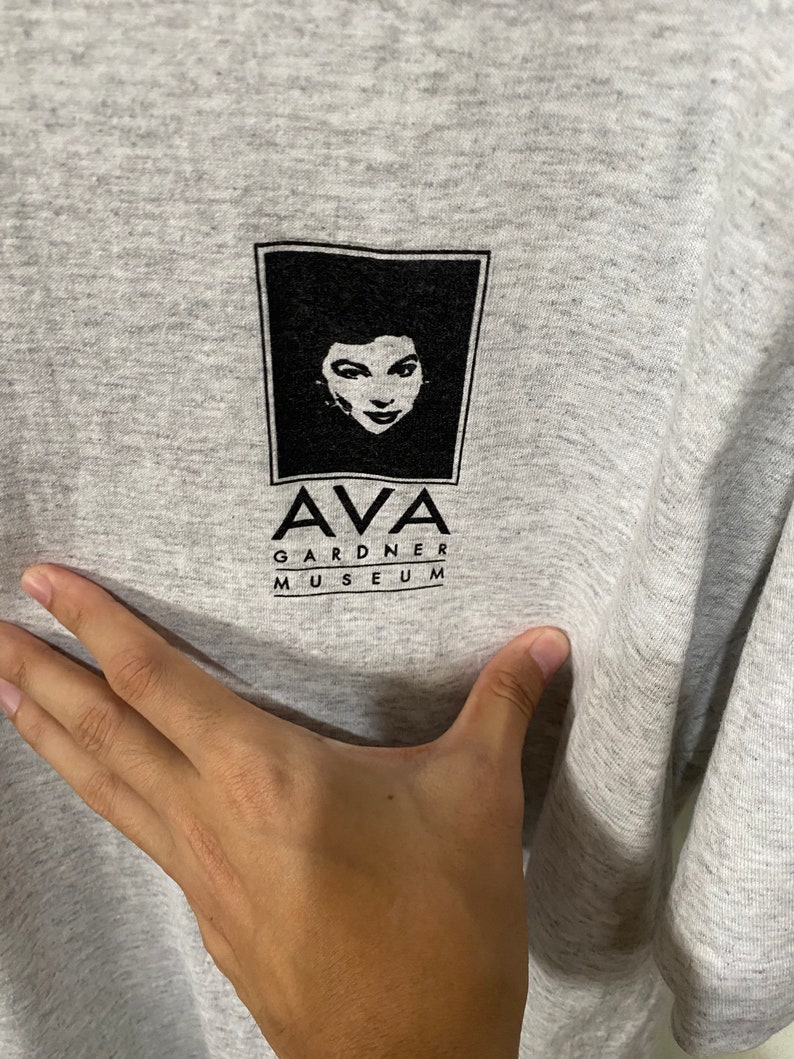 Puede incluir: Camiseta gris con un gr&aacute;fico en blanco y negro del rostro de una mujer dentro de un marco cuadrado. Debajo, el texto "AVA GARDNER MUSEUM" en negro, centrado en la camiseta.