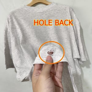 May include: A light gray t-shirt with the words "HOLE BACK" in yellow and red text. The shirt has a hole in the back, highlighted with a red circle. The shirt is hanging on a wooden hanger.