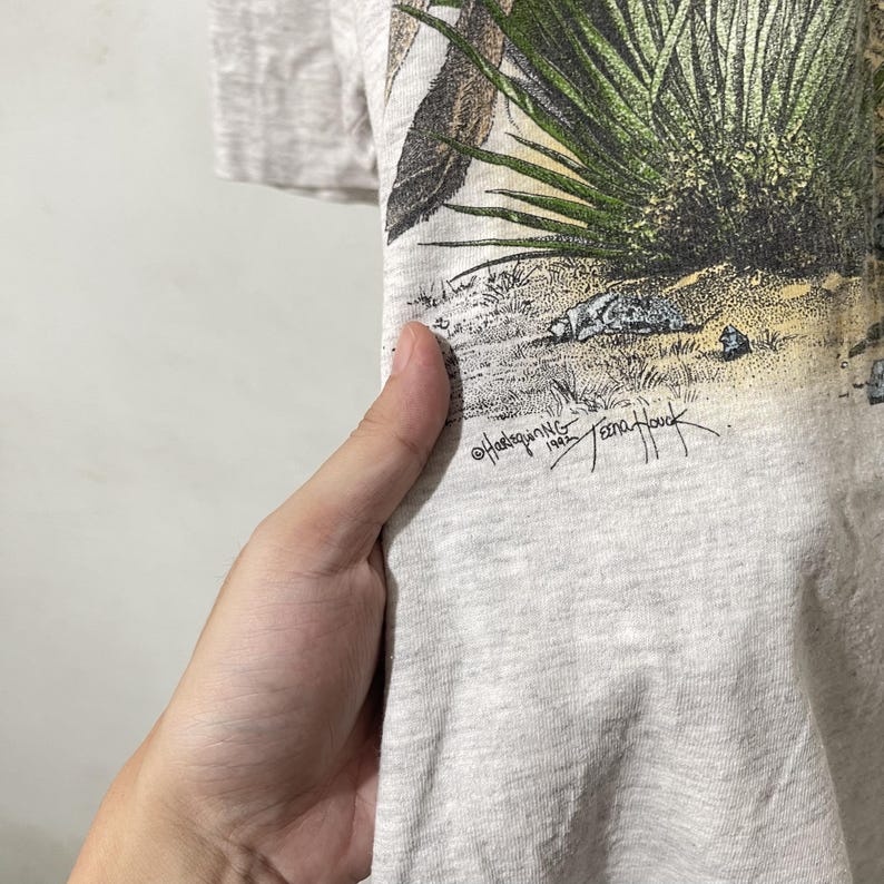 May include: Light gray t-shirt featuring a desert landscape graphic with a bird and vegetation. The shirt has a crew neck and short sleeves. The graphic includes the text "&copy;Hadsquing 1992 Teena Houck".